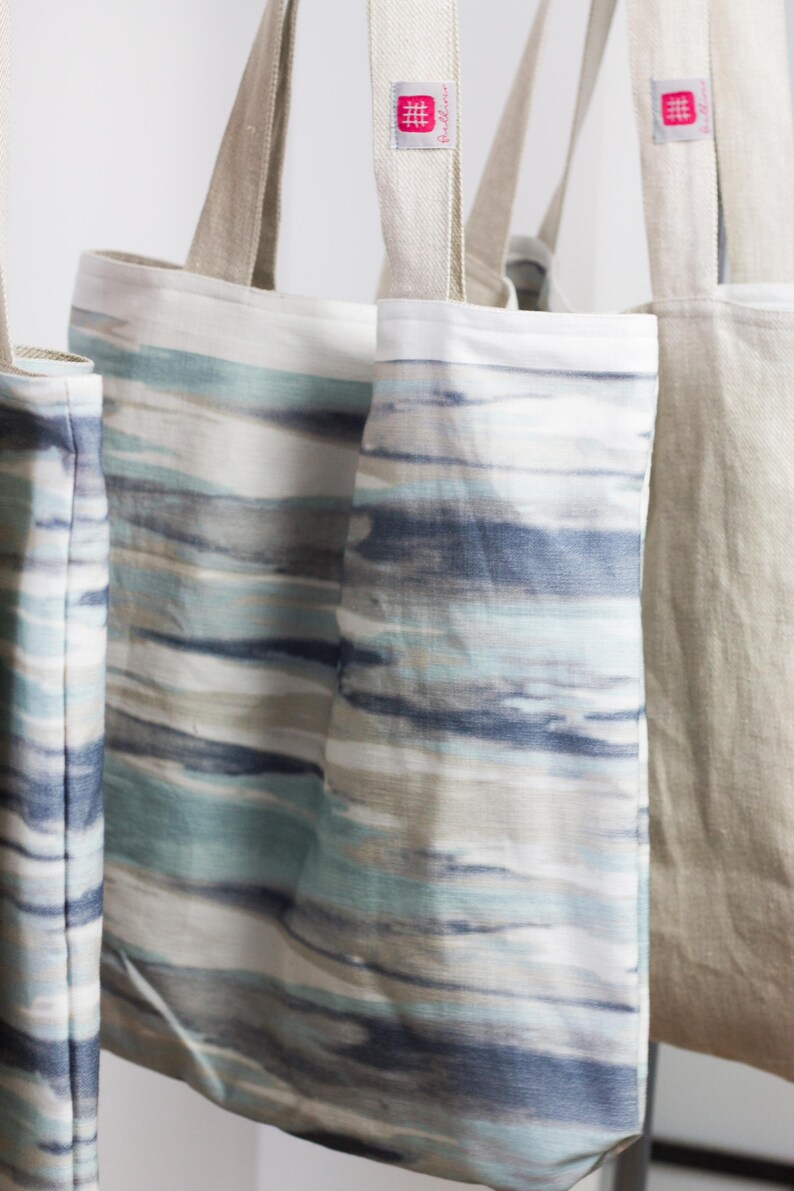 May include: Three tote bags hanging on a white wall. Two of the bags are made of natural linen and have a blue and white striped pattern. The third bag is made of natural linen and has a solid beige color. All three bags have a pink label with the word "Hollown" printed on them.