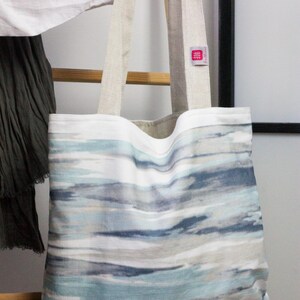 May include: A large tote bag with a white background and a blue and gray watercolor-style pattern. The bag has two long, beige canvas straps.