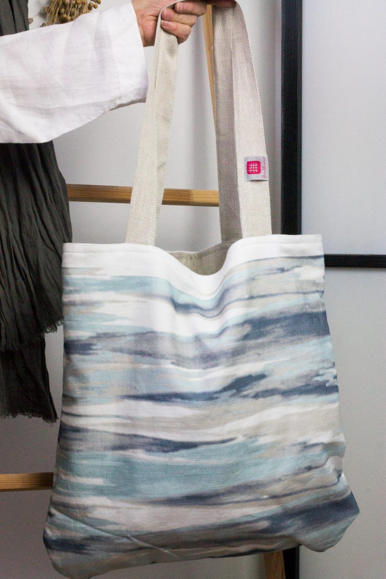 May include: A large tote bag with a white background and a blue and gray watercolor-style pattern. The bag has two long, beige canvas straps.