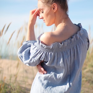 May include: A woman wearing a light blue off-the-shoulder linen dress with a ruffled neckline. She is standing in a field of tall grass with a blue sky in the background.
