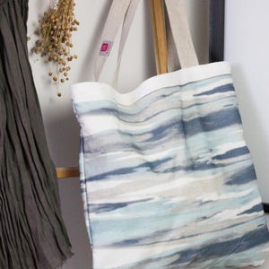 May include: A large, white canvas tote bag with blue and gray watercolor-style stripes. The bag has two long, natural linen straps.