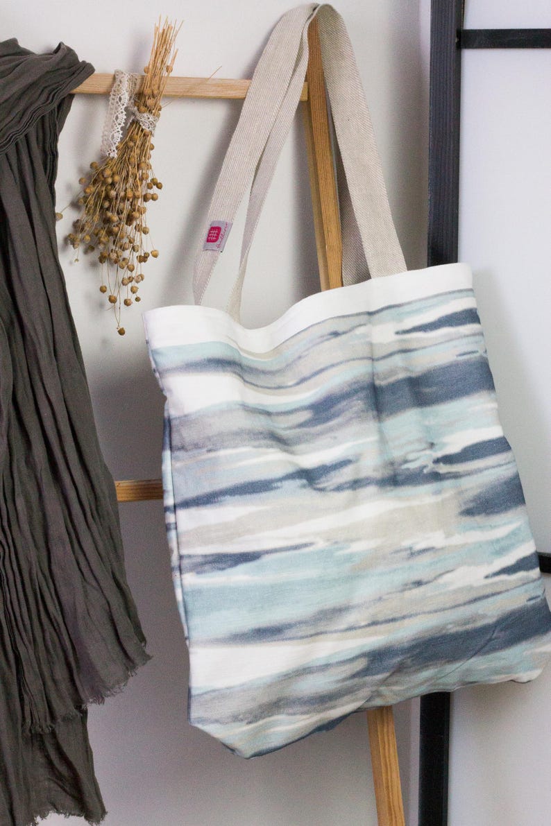 May include: A large, white canvas tote bag with blue and gray watercolor-style stripes. The bag has two long, natural linen straps.