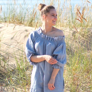 May include: A woman wearing a light blue off-the-shoulder linen dress with a ruffled neckline. She is standing in a grassy area with a sandy beach in the background.