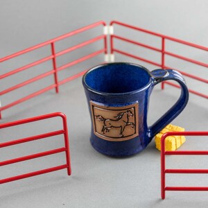 May include: A blue ceramic mug with a horse illustration, set inside a miniature red fence. The mug has a dark blue glaze and a brown rectangular emblem with a horse design. A yellow sponge is next to the mug.
