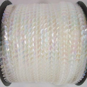May include: A spool of iridescent white sequin trim featuring a continuous strand of small, round, reflective discs. The sequins are arranged in a dense, overlapping pattern on a black plastic reel, creating a shimmering, pearlescent effect.