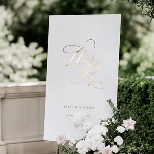 Può includere: Un cartello nuziale rettangolare bianco con un grande monogramma metallico dorato delle lettere WR. Sotto il monogramma, il testo recita WILLIAM & RACHEL e SEPT 14, 2026. Il cartello è esposto su un supporto in metallo dorato con fiori.
