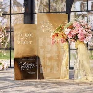 May include: Two gold mirror wedding signs with white calligraphy. The sign on the left says "You look perfect tonight." The sign on the right says "Our favorite people" and has a list of names. A bouquet of pink and yellow flowers is in a vase in front of the signs.