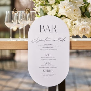 Puede incluir: Un menú de bar ovalado blanco con el texto "BAR Signature cocktails" en escritura negra. El menú enumera cócteles, cerveza, vino y licores. Copas de vino transparentes y un arreglo floral de rosas y tulipanes blancos en el fondo.