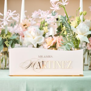 May include: A wedding reception table setting with a floral arrangement and a sign that reads "MR. & MRS. MARTINEZ" in gold lettering. The table is covered in a light green tablecloth, and the background features candles and more floral arrangements.