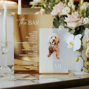 May include: A gold bar menu with a list of drinks and a smaller sign with a dog illustration and the text "Drinks ON ME". The signs are on a table with flowers and candles.