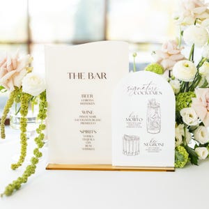 May include: A wedding bar menu with the text "THE BAR" and "signature cocktails" displayed on cream-colored cards. The menu lists beer, wine, and spirits. The cocktail menu features illustrations of drinks. Floral arrangements with pink and white roses surround the menu.