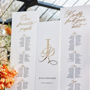 May include: White wedding seating chart with gold lettering. The chart is divided into sections labeled "One", "Two", "Three", "Four", and "Five". Each section lists names of guests. The chart also includes the couple's names, "Julia & Richard", and the date and location of the wedding, "05.31.24 | Newport, RI".