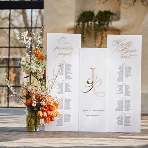 May include: White wedding seating chart with gold calligraphy. The chart is divided into three sections: "Our Favorite People", "Julia & Richard", and "Kindly Find Your Seat".