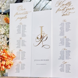 May include: White wedding seating chart with gold lettering. The chart is divided into sections labeled "One", "Two", "Three", and "Four". The top of the chart reads "Our favorite people". A separate white board with gold lettering reads "Kindly find your seat".