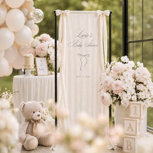 May include: A baby shower scene with a cream-colored banner that reads "Lucia's Baby Shower" and the date May 14, 2024. The scene includes a teddy bear, floral arrangements, balloons, and decorative blocks spelling "1AB".