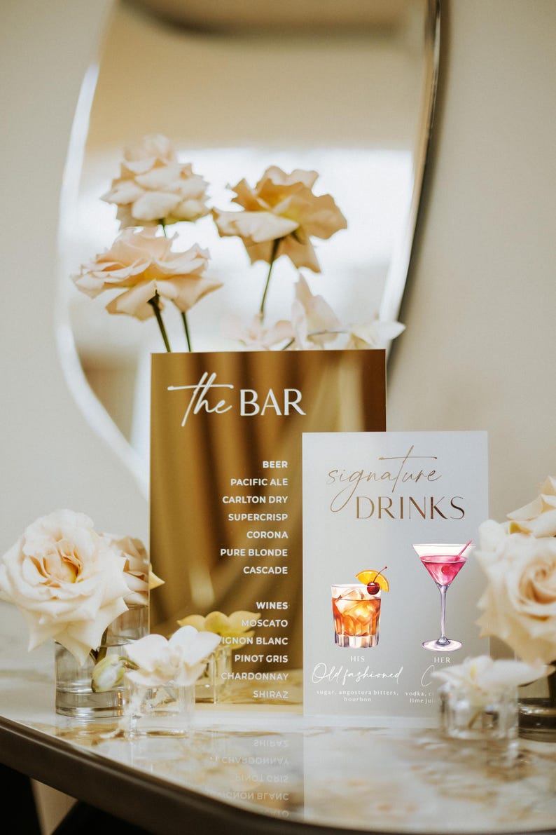 May include: A gold sign with the text "The Bar" lists beer and wine options. A second sign displays "Signature Drinks" with illustrations of cocktails. Floral arrangements and a mirror complete the elegant bar setup.