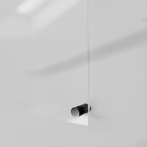 May include: A clear, rectangular glass panel with a small, cylindrical, black and silver hardware attachment. The panel is mounted on a white wall, with shadows adding depth. The minimalist design suggests a modern aesthetic.