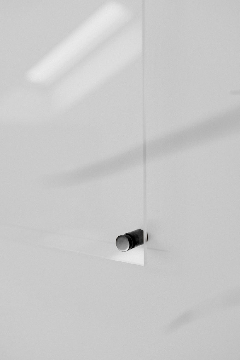May include: A clear, rectangular glass panel with a small, cylindrical, black and silver hardware attachment. The panel is mounted on a white wall, with shadows adding depth. The minimalist design suggests a modern aesthetic.