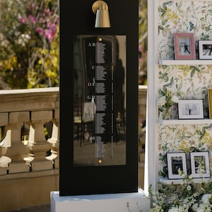 May include: A black seating chart with gold accents, featuring a list of names. A gold sconce is mounted above the chart. The chart is on a white base, with a floral arrangement to the side. A second display with photos and floral wallpaper is to the right.