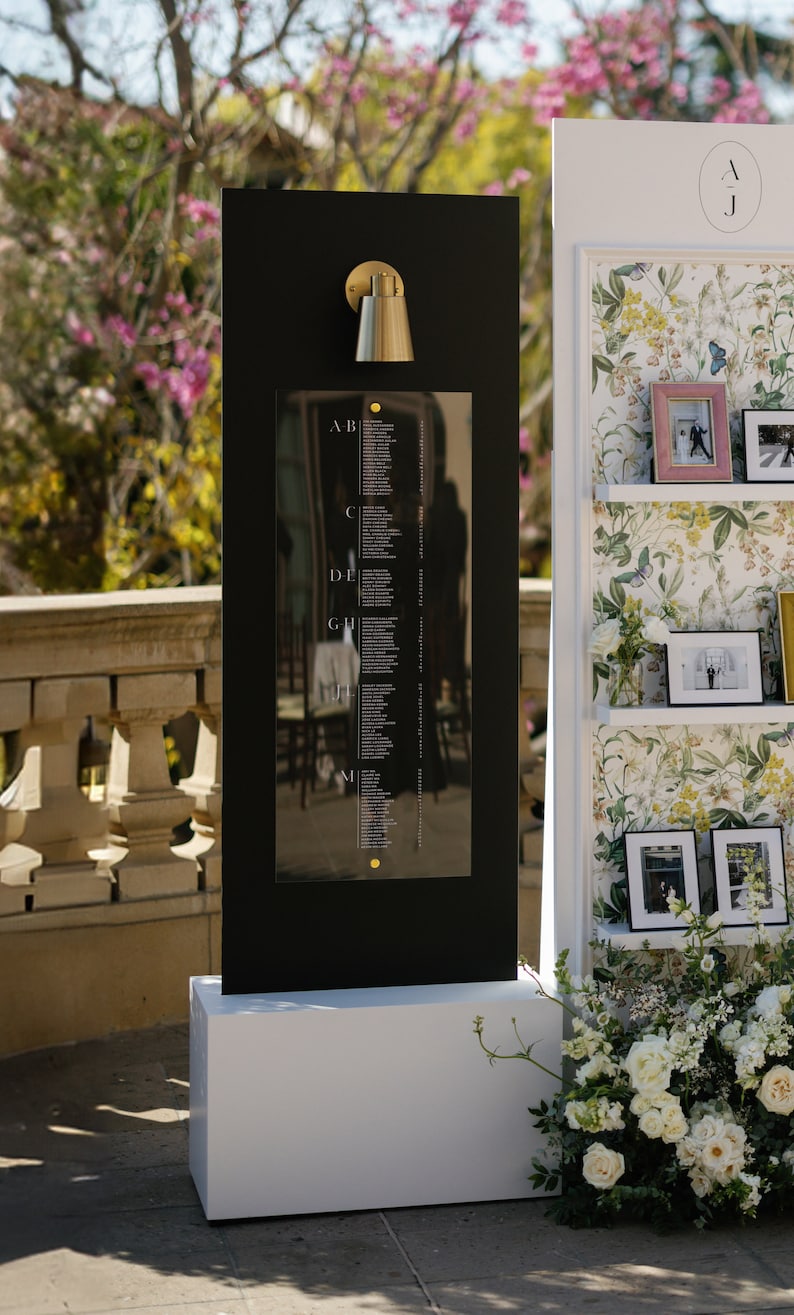 May include: A black seating chart with gold accents, featuring a list of names. A gold sconce is mounted above the chart. The chart is on a white base, with a floral arrangement to the side. A second display with photos and floral wallpaper is to the right.