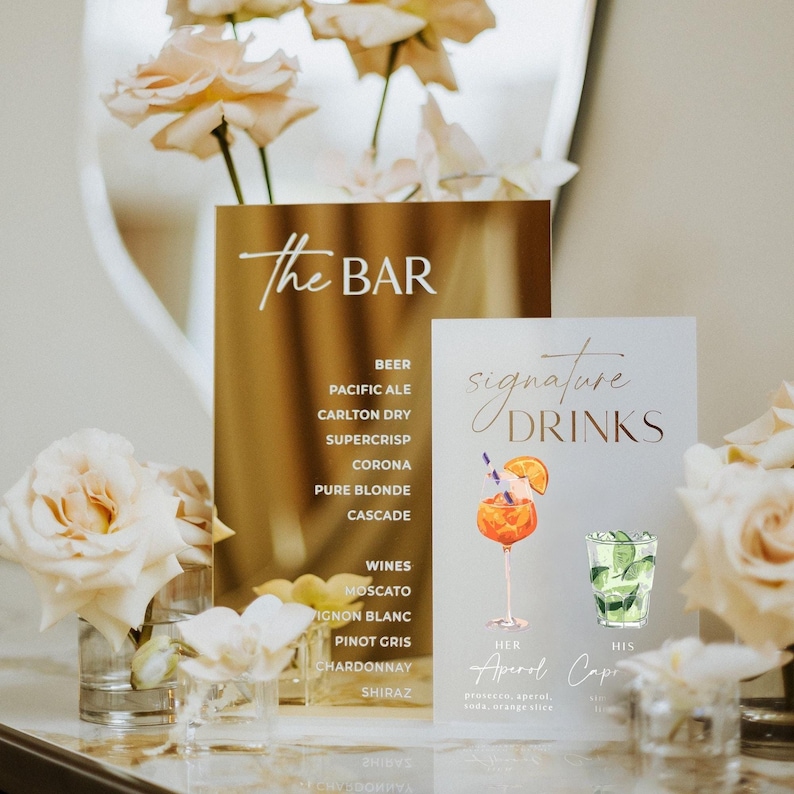 May include: Gold acrylic bar sign with the text "The BAR" and a list of beers and wines. A smaller sign displays "signature DRINKS" with illustrations of cocktails. Floral arrangements in glass vases are in the foreground.