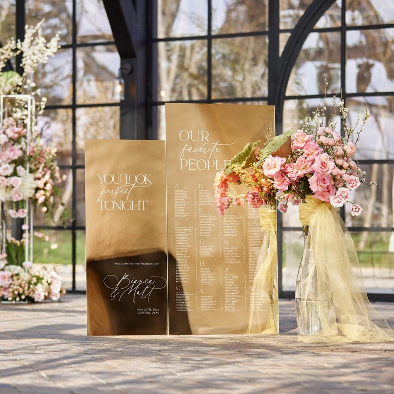 May include: Two gold mirror wedding signs with white calligraphy. The sign on the left says "You look perfect tonight." The sign on the right says "Our favorite people" and has a list of names. A bouquet of pink and yellow flowers is in a vase in front of the signs.