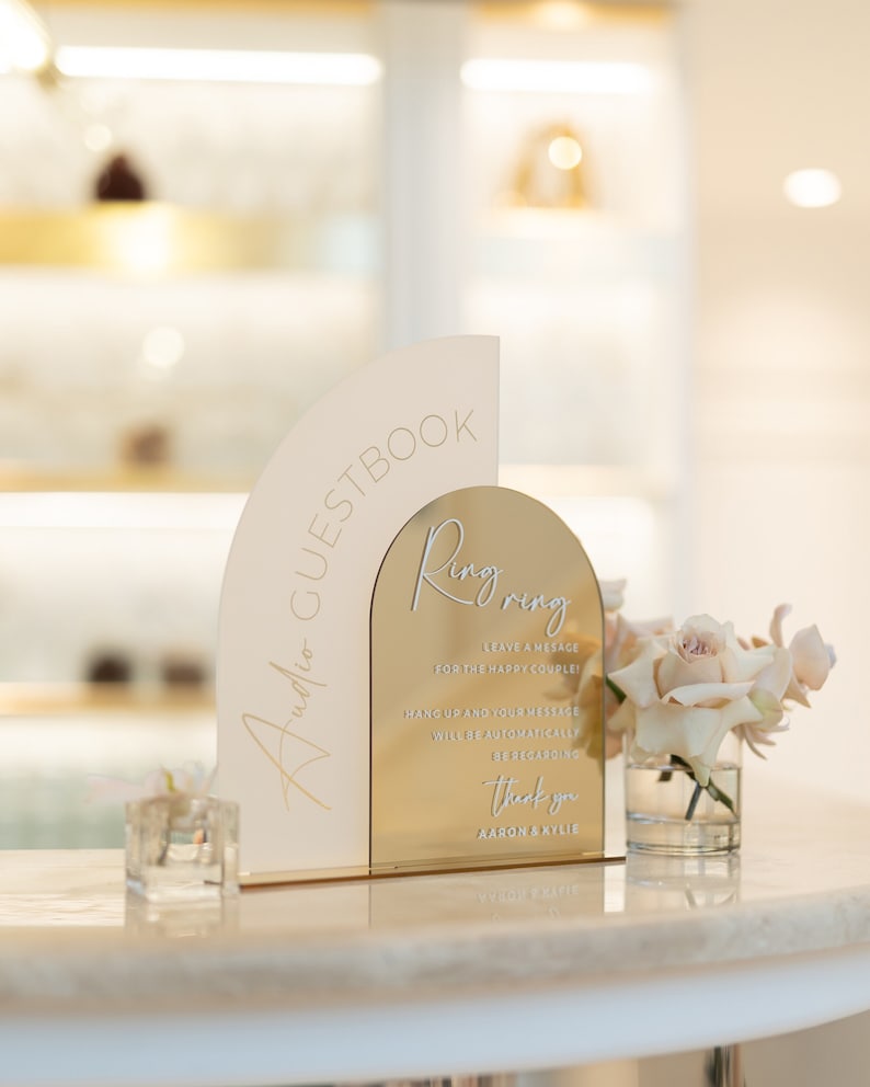May include: A gold and white arch-shaped sign with the text "Ring Ring" and instructions for guests to leave a message for the happy couple. The sign is on a white table with a vase of white roses.