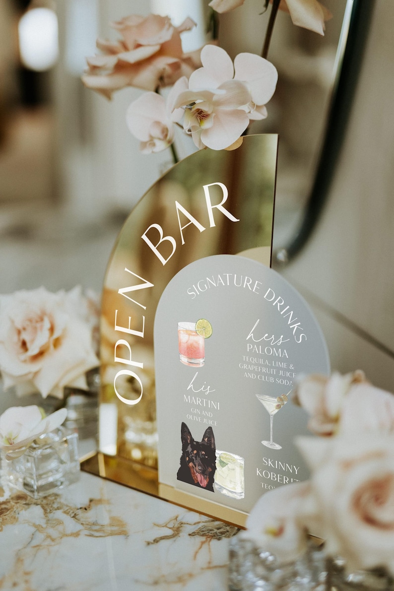 May include: A gold mirror sign with the text "Open Bar" and a white background with the text "Signature Drinks" and a list of drinks with illustrations. The drinks are "His Martini Gin and Olive Juice", "Hers Paloma Tequila, Lime & Grapefruit Juice and Club Soda", and "Skinny Koberli".