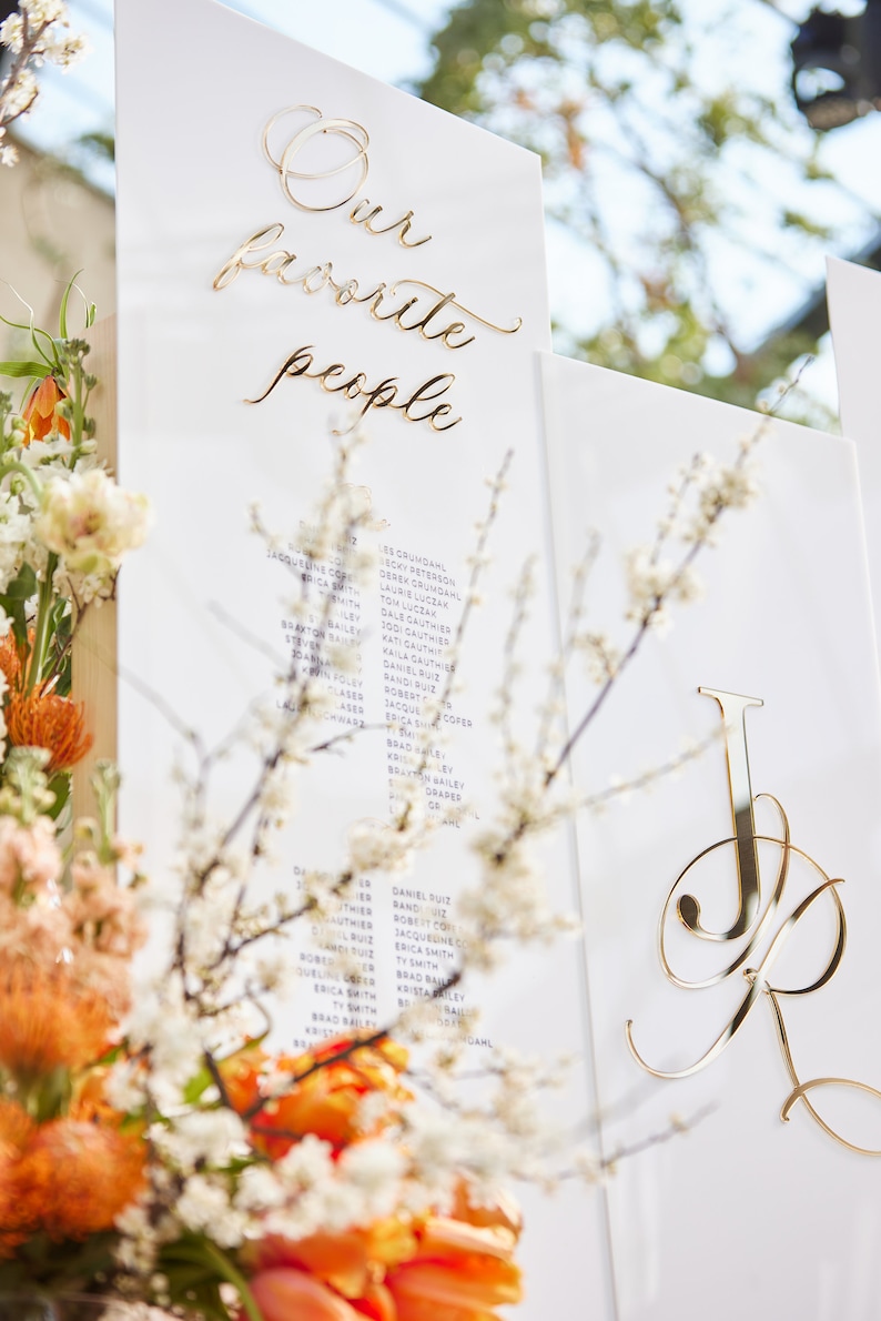 May include: A white sign with gold lettering that reads "Our favorite people" and a list of names in purple ink. The sign is leaning against a wooden surface with a bouquet of orange and white flowers in the foreground.
