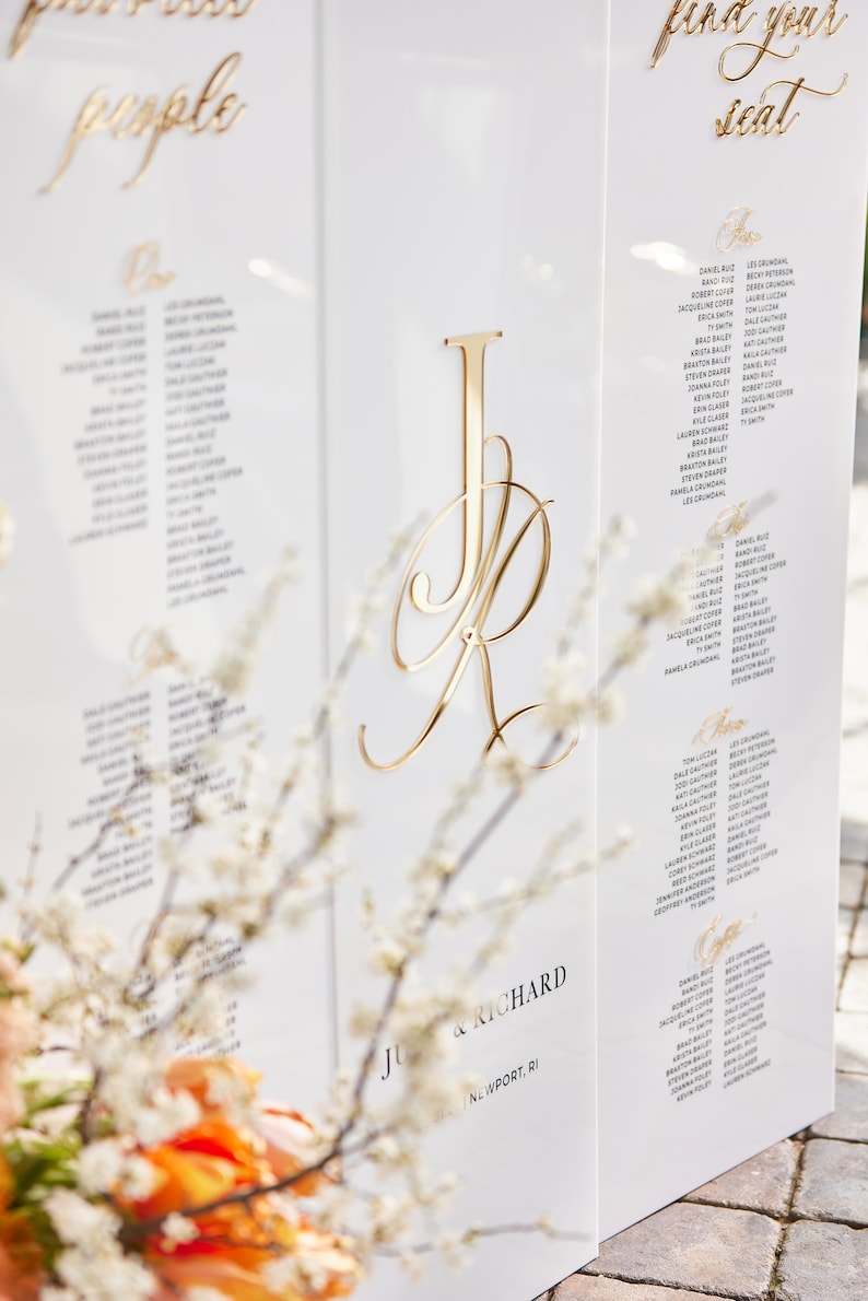 May include: A white wedding seating chart with gold lettering. The chart is divided into two sections, one for "people" and one for "find your seat". The chart features the names "Julie & Richard" and the location "Newport, RI".
