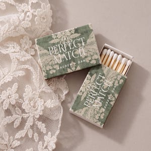 Op de afbeelding: Twee rechthoekige luciferdoosjes met een saliegroen en wit bloemenpatroon. De tekst THE PERFECT MATCH en LAUREN + DAN staat in het wit op de deksels gedrukt. Eén doosje is gesloten, het andere staat half open en toont houten lucifers met witte koppen.