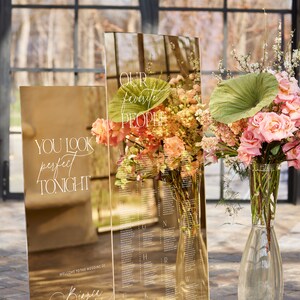 May include: A gold mirrored wedding seating chart with floral arrangements. The chart includes the text "YOU LOOK perfect TONIGHT" and "OUR favorite PEOPLE". The wedding date and location are also displayed.