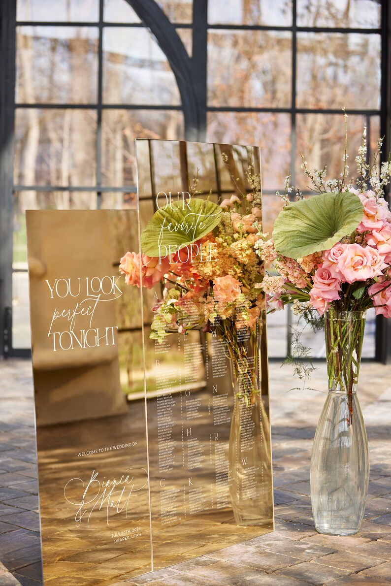 May include: A gold mirrored wedding seating chart with floral arrangements. The chart includes the text "YOU LOOK perfect TONIGHT" and "OUR favorite PEOPLE". The wedding date and location are also displayed.