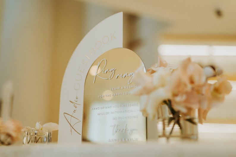 May include: A gold mirror with a white frame shaped like an arch. The mirror has the text "Ring Ring" written on it, along with instructions for guests to leave a message for the happy couple. The text "Audio Guestbook" is written on the white frame.