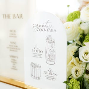 May include: A white acrylic bar menu with gold accents, featuring the text "THE BAR" and "Signature Cocktails." The menu details cocktail recipes, including a Mojito and Negroni, with illustrations. A bouquet of white flowers is in the background.