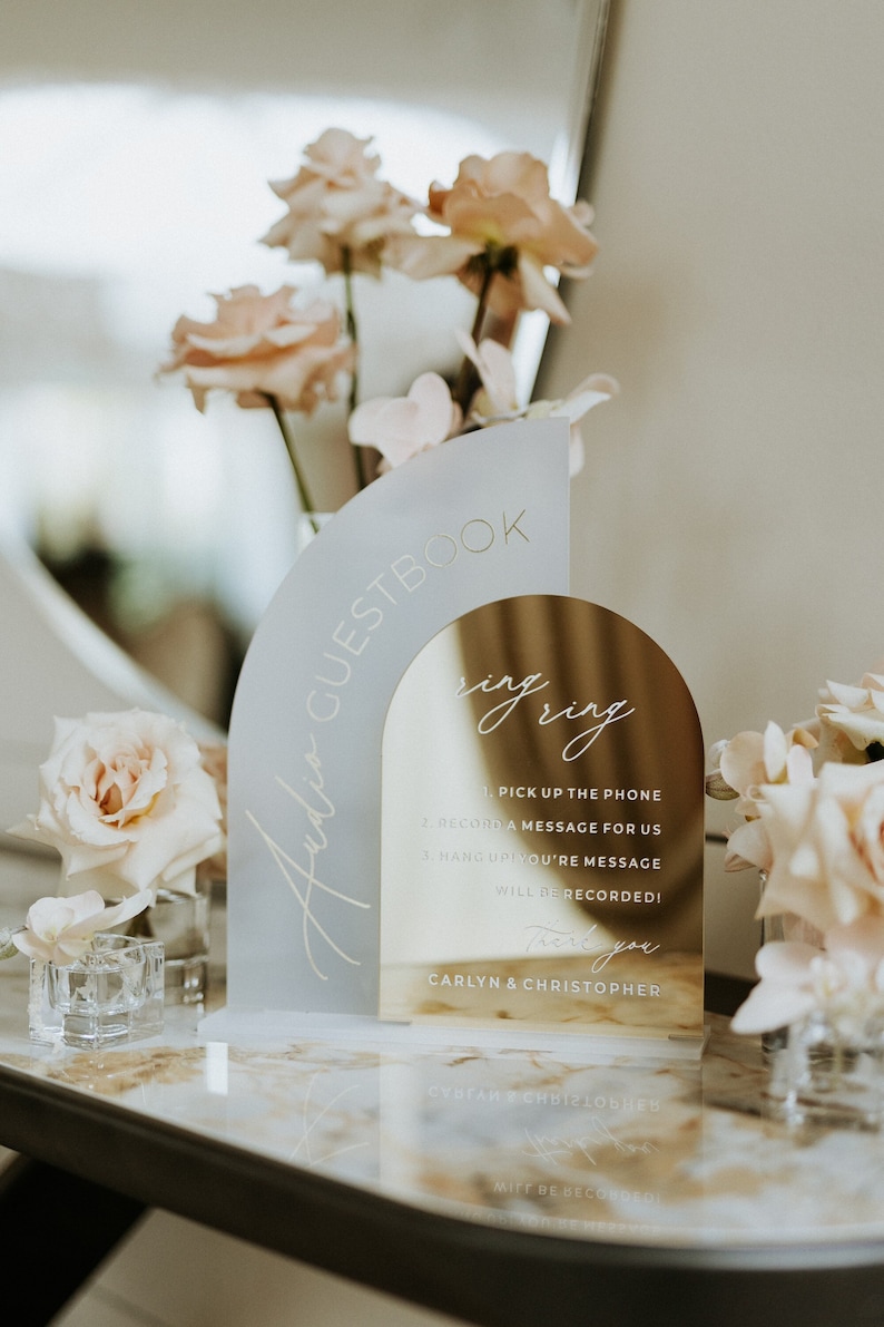 May include: A gold and white acrylic sign with the text "Audio Guestbook" and "Ring Ring" with instructions for recording a message. The sign is on a table with flowers.