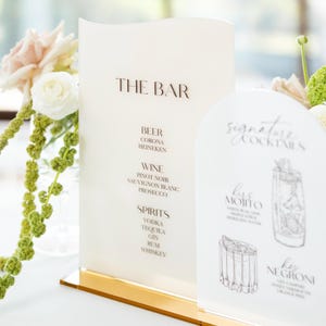 May include: Elegant bar menu with the text "THE BAR" in brown lettering. The menu lists beer, wine, and spirits. A smaller sign displays "Signature Cocktails" with illustrations of drinks. The signs are on a gold base.