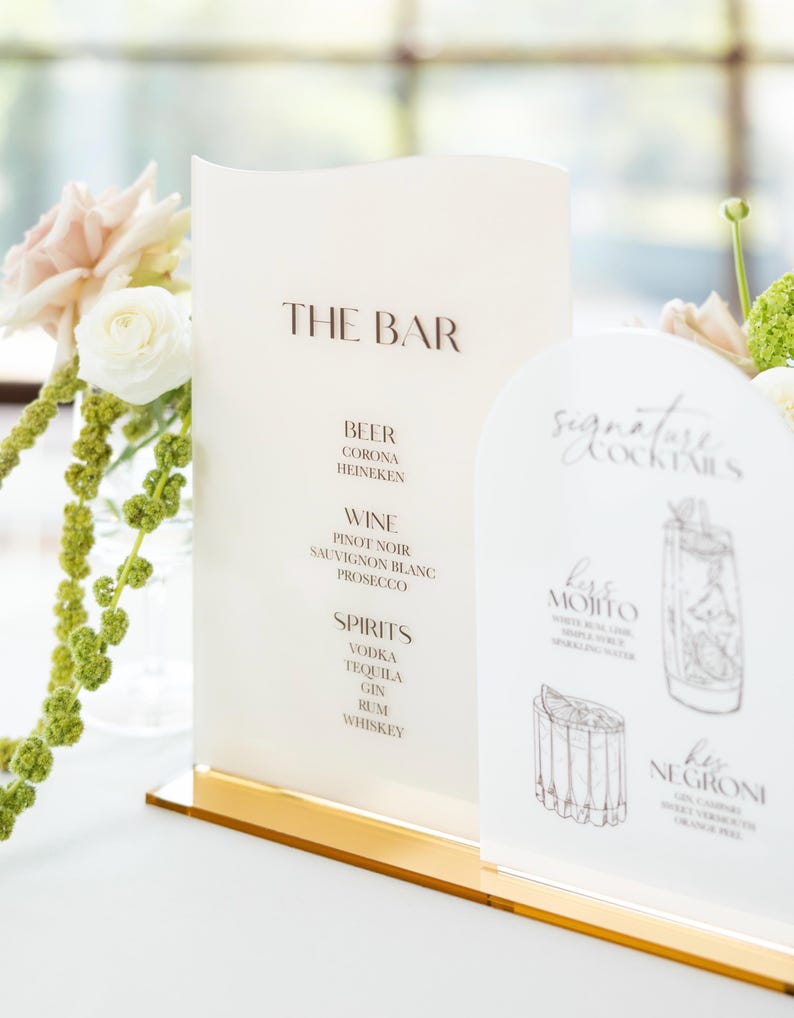 May include: Elegant bar menu with the text "THE BAR" in brown lettering. The menu lists beer, wine, and spirits. A smaller sign displays "Signature Cocktails" with illustrations of drinks. The signs are on a gold base.