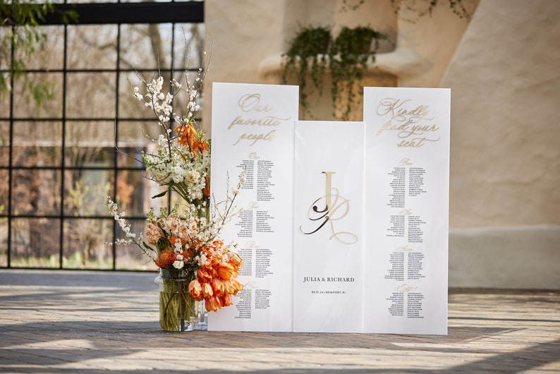 May include: White wedding seating chart with gold calligraphy. The chart is divided into three sections: "Our Favorite People", "Julia & Richard", and "Kindly Find Your Seat".