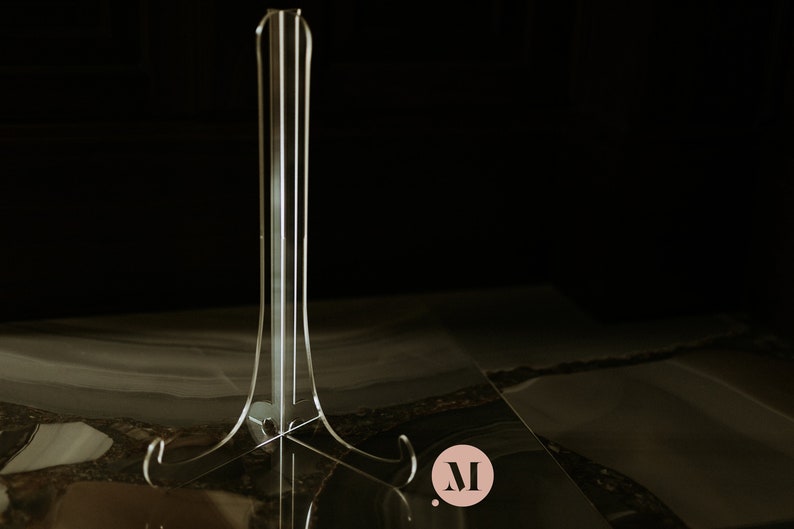 May include: A clear acrylic easel stand for displaying photos or artwork. The stand has a simple design with a tall, slender base and three legs.