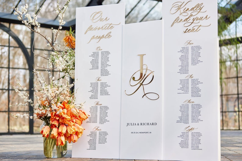 May include: White wedding seating chart with gold lettering. The chart is divided into sections labeled "One", "Two", "Three", and "Four". The top of the chart reads "Our favorite people". A separate white board with gold lettering reads "Kindly find your seat".
