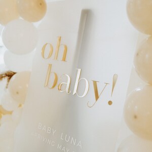 May include: A white sign with gold lettering that reads "oh baby!" surrounded by white and cream-colored balloons. The sign also includes the text "Baby Luna arriving May 2023". The sign is propped up on a dark brown stand.