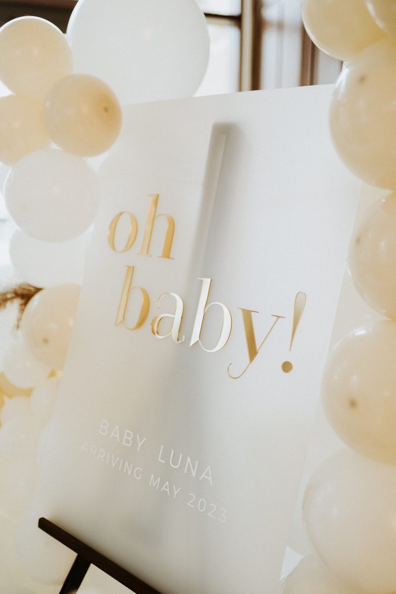 May include: A white sign with gold lettering that reads "oh baby!" surrounded by white and cream-colored balloons. The sign also includes the text "Baby Luna arriving May 2023". The sign is propped up on a dark brown stand.