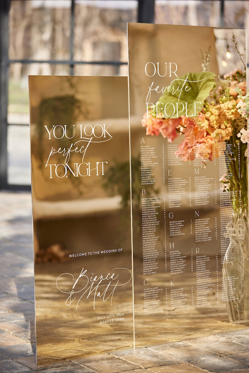 May include: Two mirrored acrylic wedding signs with white calligraphy. One sign reads "You look perfect tonight" and the other lists names. The signs are decorated with flowers and the wedding date.