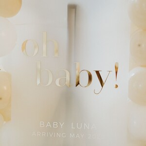 May include: A frosted acrylic sign with gold lettering that reads "oh baby!" and the name "Baby Luna" with the arrival date. The sign is surrounded by white and beige balloons, perfect for a baby shower.