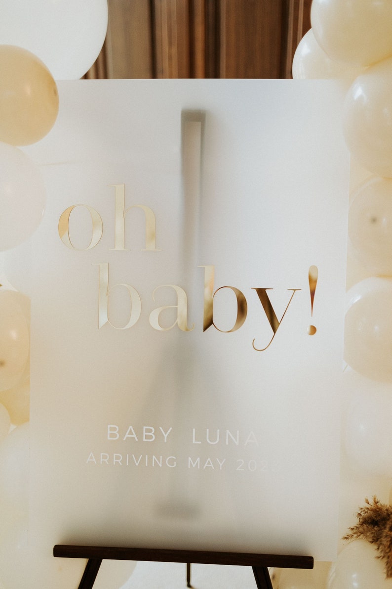 May include: A frosted acrylic sign with gold lettering that reads "oh baby!" and the name "Baby Luna" with the arrival date. The sign is surrounded by white and beige balloons, perfect for a baby shower.