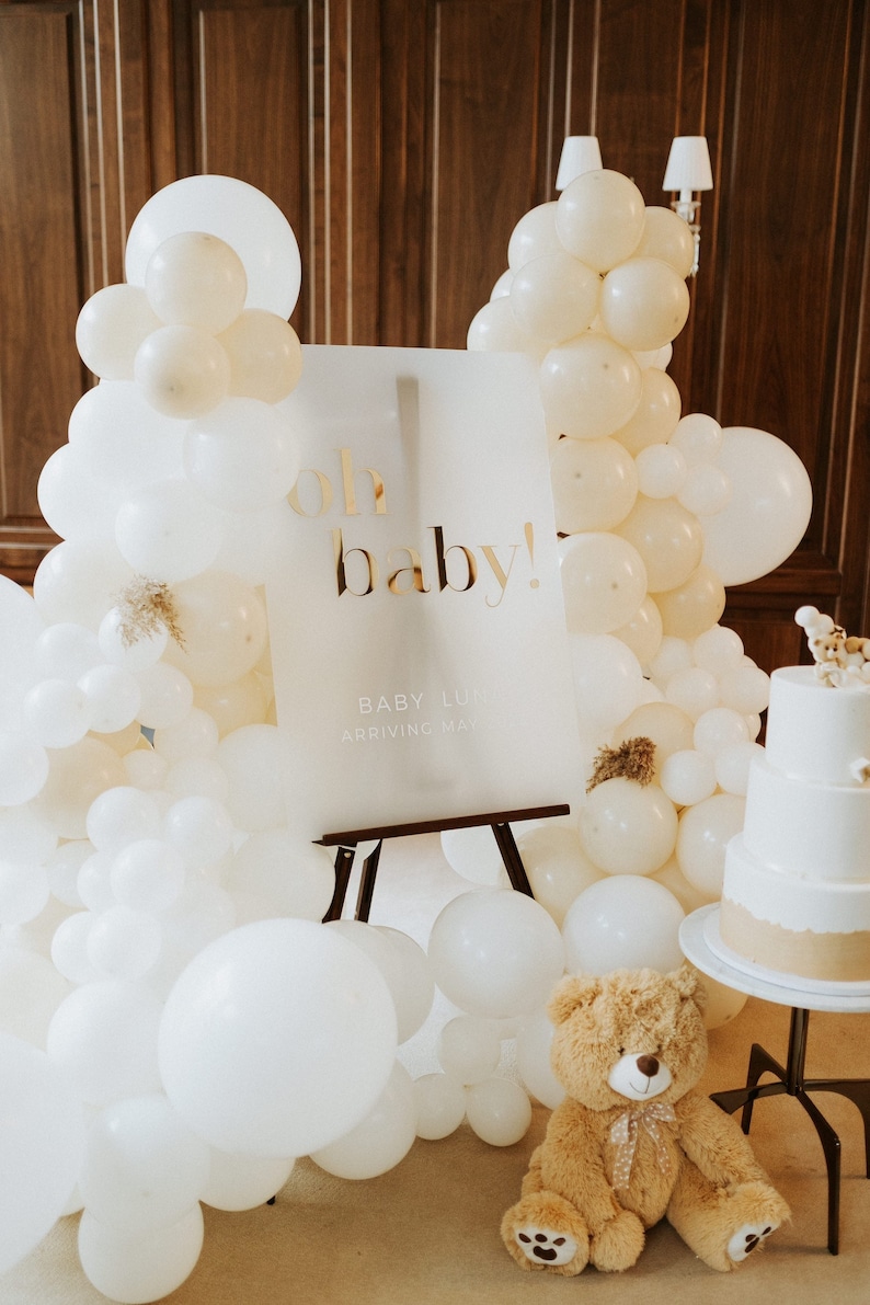 May include: A baby shower scene with a sign that reads "oh baby!" in gold lettering. The sign is surrounded by white and cream-colored balloons. A teddy bear and a tiered cake are also present, creating a festive atmosphere.