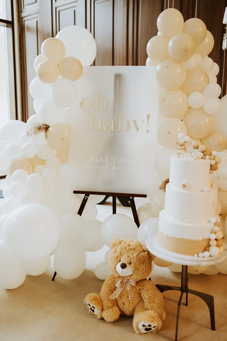 May include: A baby shower scene with a white sign that reads "oh baby!" in gold lettering. The sign is surrounded by white and cream-colored balloons. A three-tiered cake and a teddy bear are also present, creating a celebratory atmosphere.