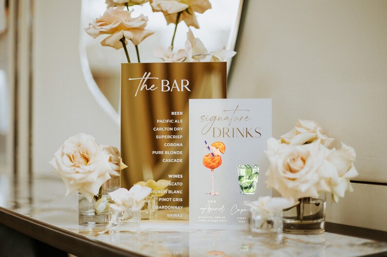 May include: A gold sign with the text "The BAR" and a list of beers and wines. A second sign displays "Signature DRINKS" with watercolor illustrations of cocktails. The signs are decorated with white roses in glass vases.