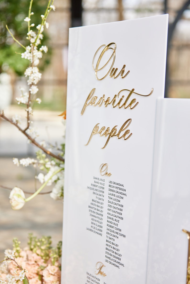 May include: A white sign with gold lettering that reads "Our favorite people" and lists names in two columns. The sign is leaning against a branch of a flowering tree.